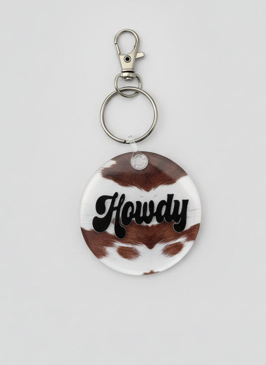 2" round keychain. Brown and white cowhide look backer with text Howdy in black.