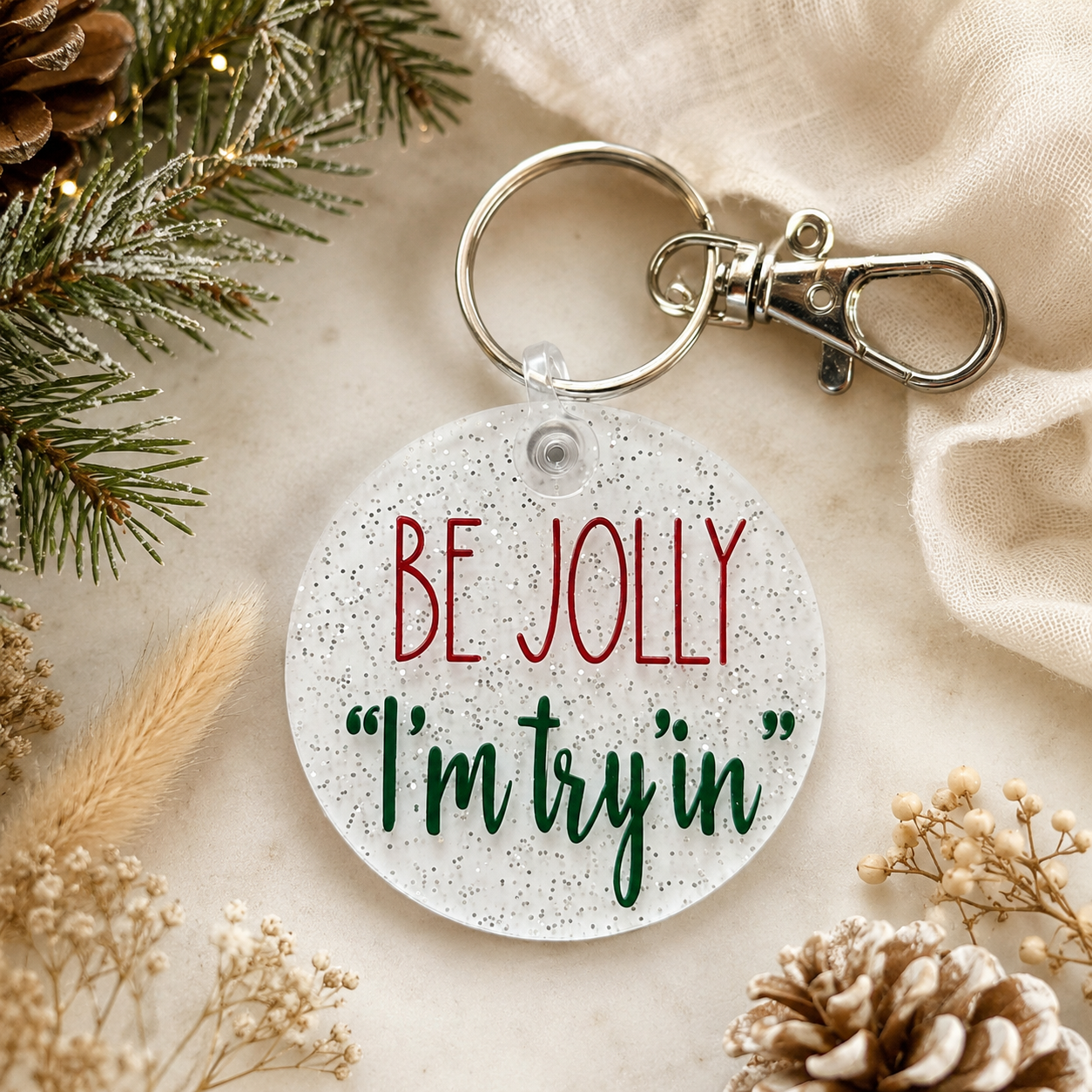 Keychain with 'Be Jolly I'm tryin'' text on a decorative background with pine branches and pinecones.
