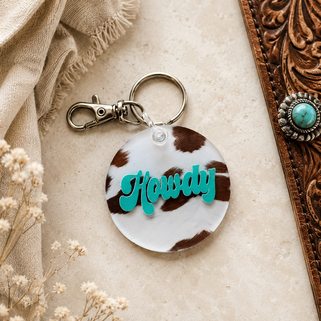 Keychain with 'Howdy' text on a brown and white patterned tag, placed on a textured surface with decorative elements.