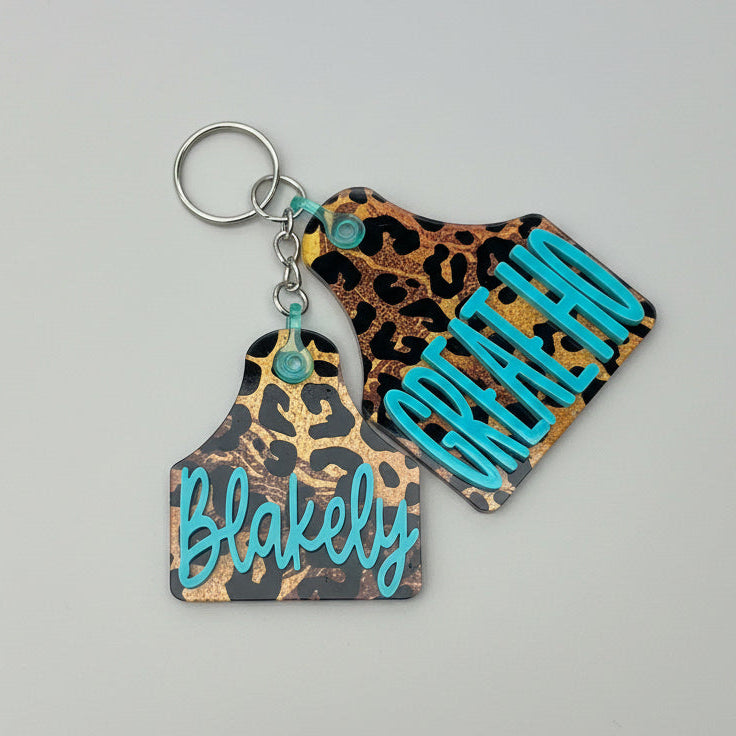 Keychain with leopard print and turquoise text held by a hand outdoors.