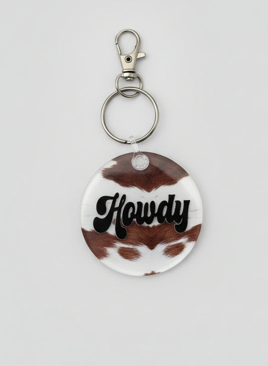2" round keychain. Brown and white cowhide look backer with text Howdy in black. 
