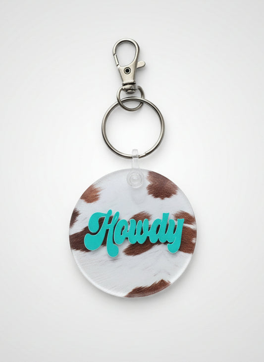 2" keychain charm. It is Brown and White Cowhide with the word Howdy in the color teal.